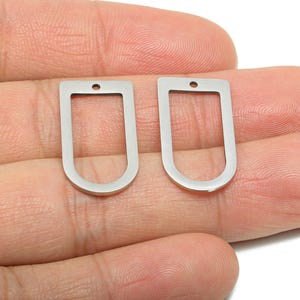 Puede incluir: Dos colgantes plateados, con marco abierto en forma de U, cada uno con un pequeño agujero en la parte superior para sujetarlos a una cadena u otro componente de joyería. Los colgantes se muestran sobre un fondo de tono de piel.