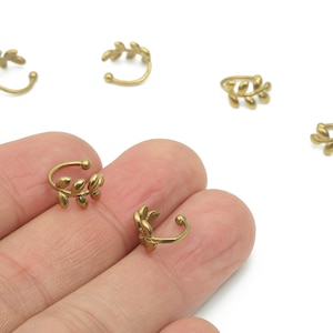 Könnte beinhalten: Goldfarbene Ohrklemmen mit Blattdesign. Mehrere Klemmen sind auf einer weißen Oberfläche verstreut, zwei davon werden von einer Hand gehalten. Die Klemmen haben eine gebogene Form, um sich dem Ohr anzupassen.
