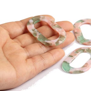 Peut inclure: Trois maillons ovales en résine avec un motif marbré rose, vert et blanc. Un maillon est tenu dans une main, tandis que deux autres sont disposés à proximité. Les maillons ont une finition lisse et brillante, pour la création de bijoux.