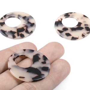 Peut inclure: Trois perles rondes et plates avec un trou central. Elles présentent un motif écaille de tortue crème, beige et marron foncé. Une perle est tenue dans une main, les deux autres sur fond blanc.