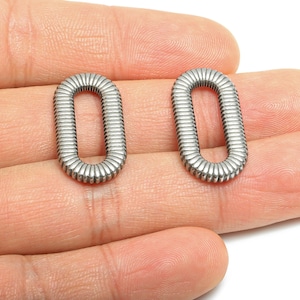 Puede incluir: Dos fornituras de joyería plateadas de forma ovalada. Cada fornitura tiene una textura acanalada y un diseño de bucle cerrado. Las fornituras se muestran en una mano, mostrando su tamaño y detalle. Adecuado para la fabricación de joyas.