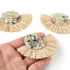 Op de afbeelding: Drie decoratieve waaier-vormige oorbellen. Elke oorbel heeft een ronde, gemarmerde, lichtgekleurde middenstuk op een waaier van natuurlijk gekleurd raffia. Eén oorbel wordt in een hand gehouden.
