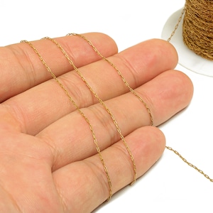 May include: A length of delicate gold-colored chain is displayed across an open palm. A spool of the same chain is visible in the background. The chain has small, rectangular links.