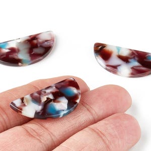 May include: Three half-moon shaped beads with a marbled pattern of burgundy, white, and teal. One bead is held in a hand, while the other two are on a white background. These beads are suitable for jewelry making.