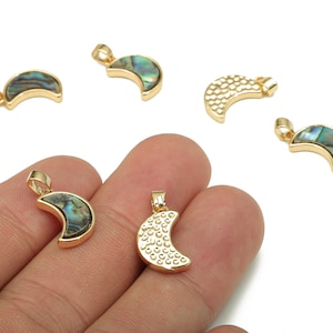 May include: Gold-toned crescent moon pendants. Some feature iridescent abalone shell inlays, while others have a textured surface. The pendants have a small loop for attaching to a chain or other jewelry component. The background is white.