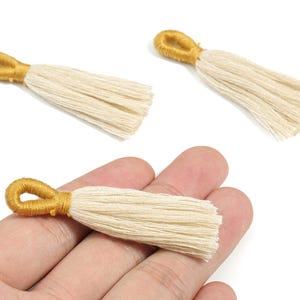 May include: Three cream-colored tassels with gold-colored loops. The tassels are made of soft, fringed material. One tassel is held in a hand. These decorative tassels are suitable for jewelry making or craft projects.