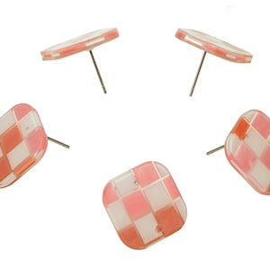 Puede incluir: Cinco pendientes cuadrados con un patrón de tablero de ajedrez en tonos rosa, blanco y naranja. Cada pendiente tiene un poste plateado. Los pendientes están sobre un fondo blanco.