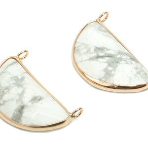 Può includere: Due ciondoli a forma di mezzaluna con una pietra marmorizzata bianca e grigia incastonata in una cornice di metallo color oro. Ogni ciondolo ha un piccolo anello nella parte superiore per il fissaggio a una catena o ad un altro componente di gioielleria. I ciondoli sono esposti su sfondo bianco.