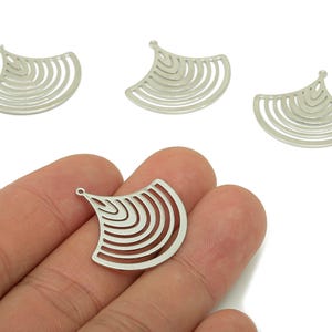 May include: Silver-colored metal fan-shaped charms with a series of curved lines. The charms have a small loop at the top for attaching to jewelry. The image shows several charms, with one held in a hand.