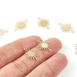 Puede incluir: Encantos dorados en forma de sol con superficies texturizadas y pequeños bucles para la fabricación de joyas. Varios amuletos están esparcidos sobre un fondo blanco, con dos sostenidos en una mano.