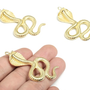 May include: Gold-coloured cobra-shaped pendants. The pendants have textured scales and a loop at the top for stringing. One pendant is held in a hand. The cobra is a popular symbol in jewellery.