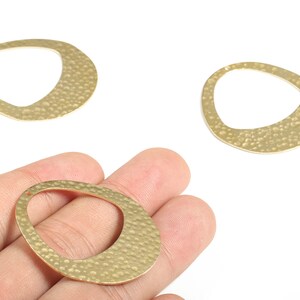 May include: Three gold-colored, open-center oval pendants with a hammered texture. One pendant is held in a hand, while two others are displayed on a white background. These pendants are suitable for jewelry making.