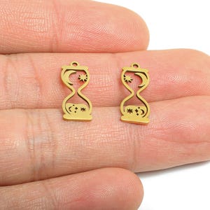 May include: Two gold-coloured hourglass charms. Each charm features a crescent moon and star design at the top, with stars at the bottom. The charms are held in a hand.