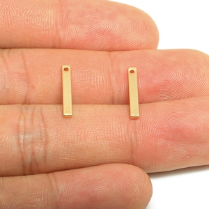May include: Two small, gold-coloured rectangular charms with a hole at the top. These charms are shown against a skin-toned background. The charms are likely for jewellery making.