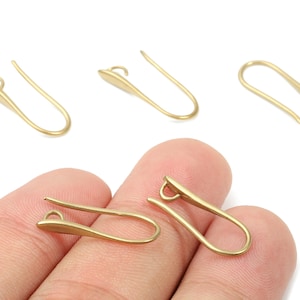 May include: Several gold-colored ear hooks are displayed against a white background. Two hooks are held by a hand. The hooks have a curved design with a small loop at the top and a flat, elongated bottom.