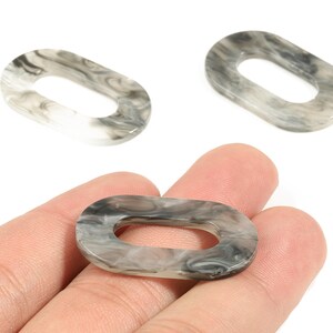 May include: Three oval-shaped acrylic links with a marbled gray and white pattern. One link is held in a hand, while the other two are positioned above. These links are suitable for jewelry making or craft projects.
