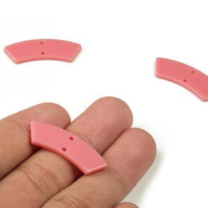 Könnte beinhalten: Drei rosa, gebogene Knöpfe mit je zwei Löchern. Ein Knopf wird in einer Hand gehalten, während die anderen beiden auf weißem Hintergrund verstreut sind. Die Knöpfe haben eine glatte, glänzende Oberfläche und eine leicht gewölbte Form, geeignet für Nähprojekte.
