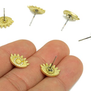 May include: Gold-coloured stud earrings in an eye shape with a sunburst design. The centre of the eye is encrusted with small, clear crystals. The earrings are shown on a white background.