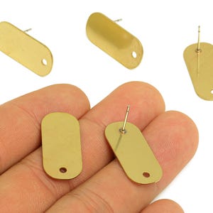 Può includere: Perni per orecchini rettangolari color oro con un piccolo foro nella parte inferiore. Gli orecchini sono mostrati in varie posizioni, alcuni con il perno attaccato. Gli orecchini sono un design semplice e minimalista.