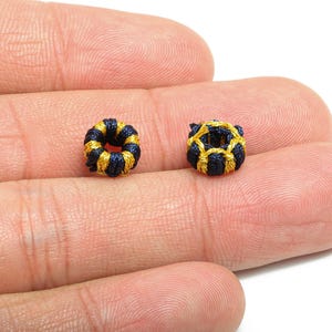 Könnte beinhalten: Zwei kleine, dekorative, handgenähte Perlen. Die Perlen sind marineblau und goldfarben, mit einer kreisförmigen Form. Eine Perle ist ein fester Ring, die andere ein käfigartiges Design. Die Perlen werden auf einer Hand gezeigt.