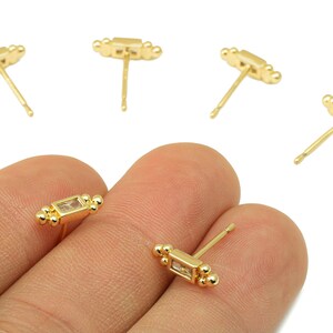 May include: Gold-colored stud earrings with a rectangular, clear stone and small ball accents. The earrings are displayed on a hand, with several others scattered in the background. The earrings have a post backing.