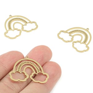 Könnte beinhalten: Goldfarbene Metall-Regenbogen-Anhänger mit Wolkenakzenten. Die Anhänger haben ein Ausschnitt-Design mit einer kleinen Schlaufe oben zum Befestigen an Schmuck oder anderen Bastelarbeiten. Drei Anhänger sind sichtbar, einer davon wird in einer Hand gehalten.