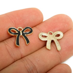 Op de afbeelding: Twee strikvormige bedels. Eén is zwart met een gouden omtrek, de andere is goudkleurig met een textuuroppervlak. Beide hebben een kleine lus aan de bovenkant om aan sieraden of andere knutselwerken te bevestigen. Ideaal voor doe-het-zelf projecten.