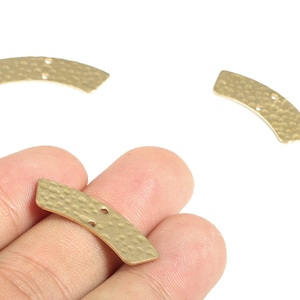 Puede incluir: Tres componentes de joyería de metal dorado, curvados, con una textura martillada. Cada pieza tiene dos pequeños agujeros. Una pieza se sostiene en una mano, las otras dos sobre un fondo blanco.