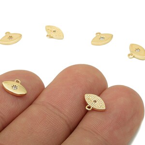 May include: Gold-colored oval charms with a starburst design. The charms have a small loop at the top for attaching to jewelry. Several charms are scattered on a white background, with some held by a person's fingers.