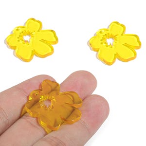 Pode incluir: Três pingentes de acrílico em forma de flor amarela. Dois pingentes estão em um fundo branco e um é segurado em uma mão. Os pingentes têm um buraco no centro e são translúcidos.