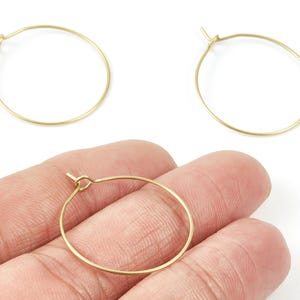 May include: Three gold-colored hoop earrings. Two are displayed separately, and one is held in a hand. The earrings are simple, circular, and made of thin metal wire. They are designed to be lightweight and easy to wear.