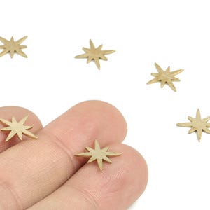 May include: Small, gold-colored star charms with eight points are scattered on a white background. Some of the charms are held between fingers. These charms are suitable for jewelry making and other craft projects.
