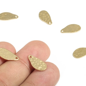 Peut inclure: Gros plan de plusieurs petits breloques métalliques dorées en forme de goutte d'eau. Chaque breloque a une surface texturée et un petit trou en haut pour l'attacher à des bijoux ou à d'autres créations. Les breloques sont éparpillées sur un fond blanc.
