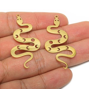 May include: Two gold-coloured snake-shaped charms. Each charm has a head with two small dots, a curved body with crescent moon cutouts, and a curled tail. The charms are made of a shiny, metallic material.