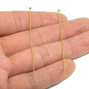Peut inclure: Deux épingles à tête dorées, chacune avec une petite boule au sommet, sont tenues dans une main. Les épingles sont droites et fines, et les boules sont lisses et rondes. Ces épingles sont probablement utilisées pour la fabrication de bijoux ou d'autres projets d'artisanat.