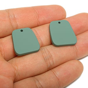 May include: Two sage green, rectangular pendants with rounded corners and a small hole at the top. The pendants are displayed on a hand, showcasing their smooth, matte finish. Ideal for jewelry making.