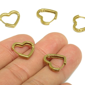 Puede incluir: Pendientes de aro dorados en forma de corazón. Los pendientes están hechos de un metal brillante y tienen un diseño simple y elegante. Los pendientes se muestran sobre un fondo blanco.