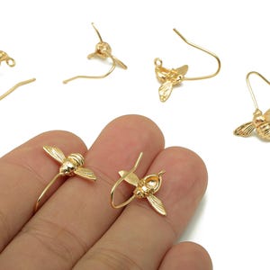 Puede incluir: Pendientes dorados con forma de abeja con alas y cuerpos detallados. Los pendientes tienen un diseño de gancho para facilitar su uso. La imagen muestra varios pares de pendientes, algunos de ellos sostenidos en una mano.