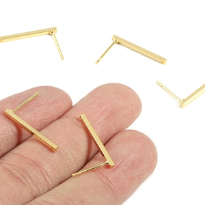 May include: Gold-colored bar stud earrings. The earrings have a simple, minimalist design, with a straight bar shape and a post for insertion. The earrings are shown against a white background.