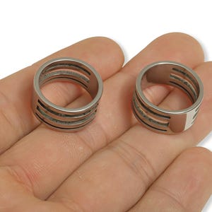 Op de afbeelding: Twee zilverkleurige ringen met een modern design. Elke ring heeft een reeks horizontale uitsparingen, waardoor een gelaagd effect ontstaat. De ringen worden op een hand getoond, waarbij hun metallic glans en details worden getoond. Sieraden.