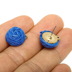 May include: A pair of blue woven stud earrings. Each earring has a round, woven design in a vibrant blue colour, attached to a gold-coloured metal backing. The earrings are shown on a person's hand.