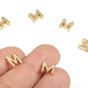 Può includere: Ciondoli a forma di lettera "M" in metallo color oro. I ciondoli sono piccoli, con un semplice carattere squadrato. Diversi sono sparsi su uno sfondo bianco, con due tenuti tra le dita di una persona. Questi ciondoli sono probabilmente per la creazione di gioielli o l'artigianato.