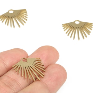 Pode incluir: Pingentes de metal dourado em forma de leque com um pequeno laço na parte superior para fazer joias. Os pingentes têm um design de explosão solar com várias linhas retas irradiando para fora. A imagem mostra três pingentes, um deles na mão.