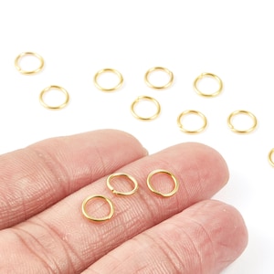 May include: Close-up of small, gold-colored jump rings. Several rings are scattered on a white surface, with a few held between fingers. These jewelry findings are used for connecting jewelry components.