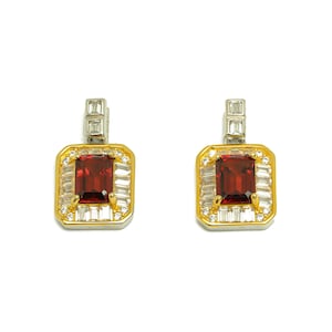 May include: A pair of rectangular earrings with a gold and silver setting. Each earring features a large, rectangular, deep red gemstone at the center, surrounded by smaller, clear gemstones. The earrings have a rectangular top.