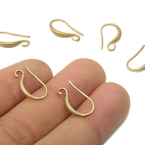 Può includere: Diversi ganci per orecchini color oro sono esposti su sfondo bianco. Due ganci sono tenuti da una mano. I ganci hanno un design curvo con un motivo strutturato nella parte inferiore. Per la creazione di gioielli.