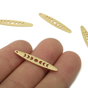May include: Gold-colored metal bar charms with a crescent moon phase design. Each charm is elongated with a series of crescent moon cutouts and a small hole at one end. The charms are shown against a white background.