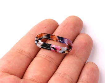 May include: An oval-shaped link with a tortoiseshell pattern in shades of brown, black, pink, and white. The link has a hollow center and is held in a hand against a white background. This decorative link is suitable for jewelry making.