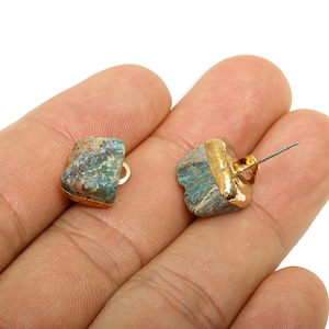 May include: A pair of stud earrings featuring rough-cut, teal-colored stones with gold-colored metal accents. The earrings have a post and backing for secure wear. The natural stone has a unique, textured appearance.