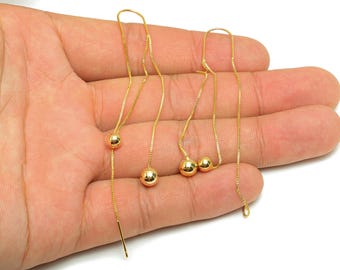 May include: Gold-colored threader earrings with small, round beads. The earrings are displayed against a light background. The design features a delicate chain that hangs down, with a small ball at the end of each chain.